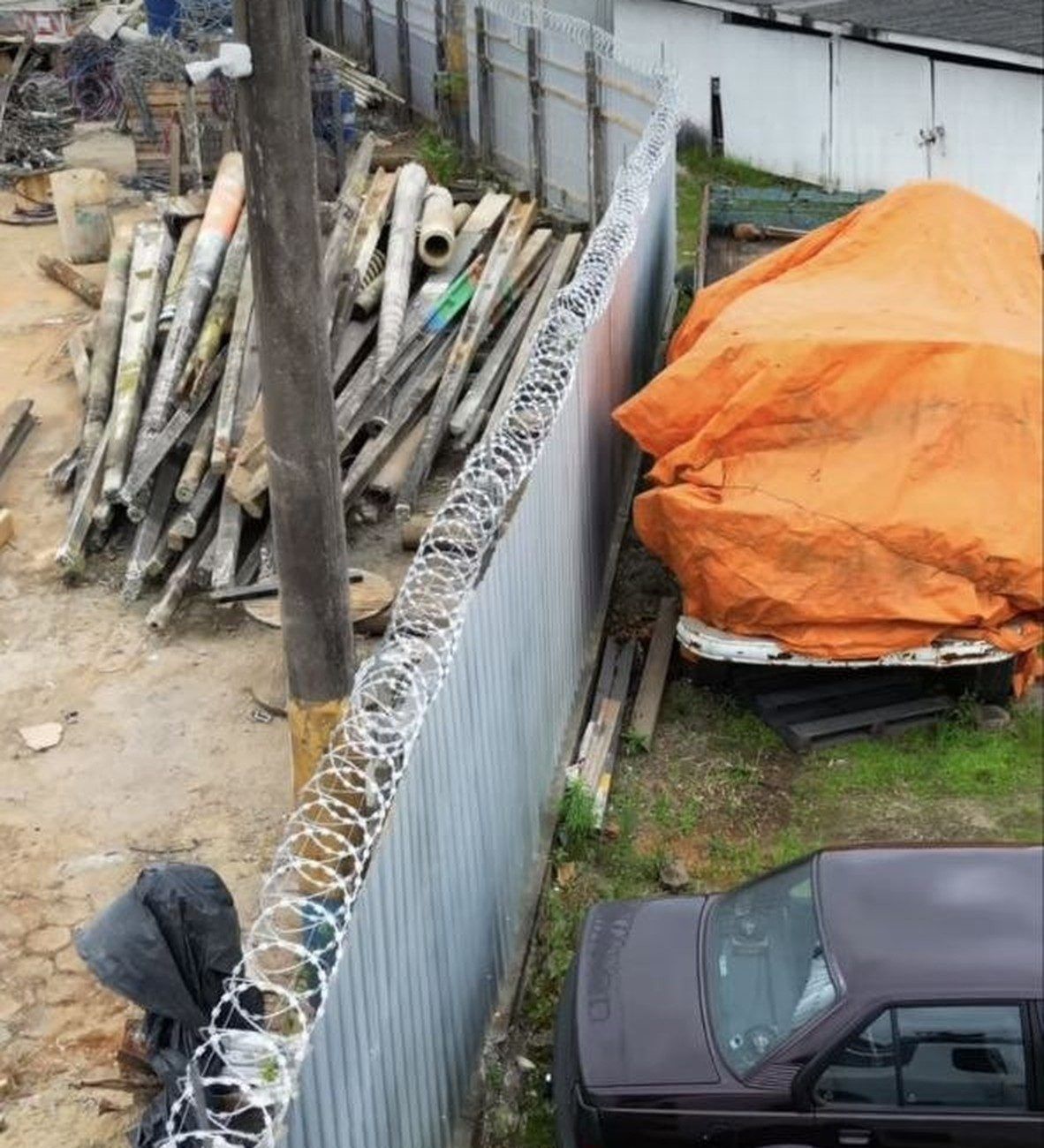 Concertina em espiral instalada sobre um muro de telha metálica, vista de cima, separando um pátio de obra de um estacionamento